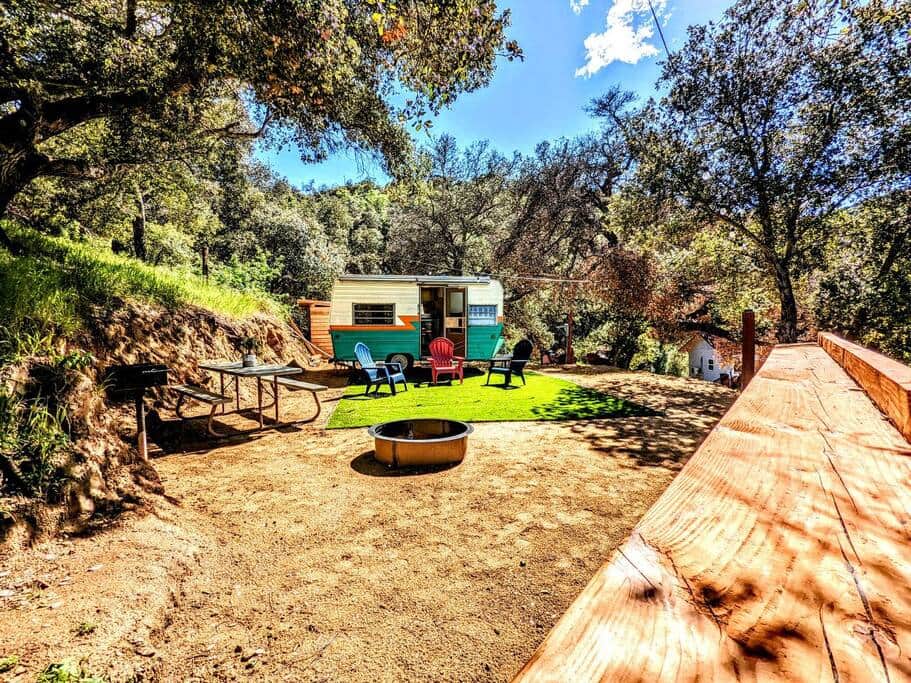 Vintage teal and cream travel trailer parked in an oak grove with Adirondack chairs around a fire pit